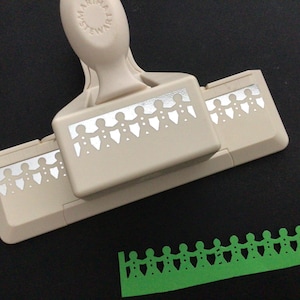 May include: A beige paper punch with a white border design of people holding hands. The punch is on a black surface with a green paper border design of people holding hands.