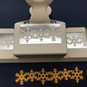 May include: A white snowflake punch with a silver snowflake design. The punch is used to create snowflake shapes from paper. A row of yellow paper snowflakes is shown below the punch.