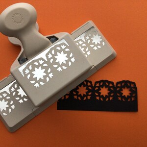 May include: A beige paper punch with a decorative border design. The punch has three different border designs. A black paper strip with the same border design is shown below the punch.