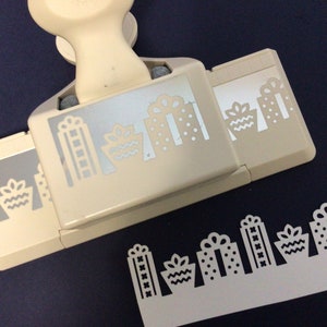 May include: A beige paper punch with a handle and a lever. The punch creates a border of small gift boxes with bows, polka dots, and wavy lines. The punch is on a dark blue surface.
