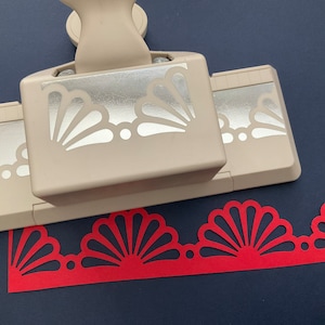 May include: A beige paper punch with a silver design of a scallop shell border. The punch is used to create a red paper border with a scallop shell design.
