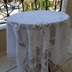 May include: A round white crochet tablecloth featuring an intricate openwork pattern with floral motifs and a scalloped edge with hanging fringe. The textile is draped over a circular table, displaying a detailed needlework design throughout.