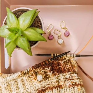 Peut inclure: Gros plan sur un projet de tricot en laine brune et blanche. Le projet est sur un plateau rose avec une plante succulente et trois marqueurs de point en or et en cristal.