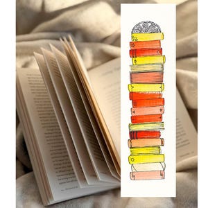 Op de afbeelding: Een open boek met uitwaaierende pagina's, naast een aquarel bladwijzer. De bladwijzer toont een stapel kleurrijke boeken in geel, oranje en rood, bekroond met een citroenschijf ontwerp.