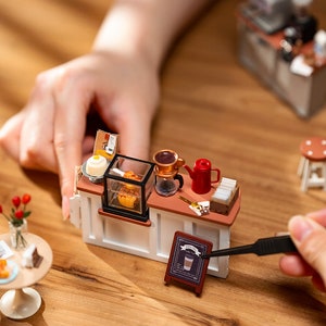 May include: A miniature coffee shop playset with a white counter, a red coffee pot, a glass display case with pastries, and a brown sign that says "Coffee Shop".