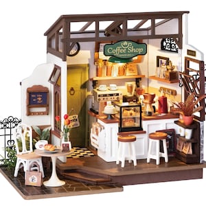May include: A miniature dollhouse coffee shop with a white exterior, brown roof, and a sign that reads "Coffee Shop Since 1903". The shop has a small patio with a table and chairs, and a window with a sign that reads "We love what we do".