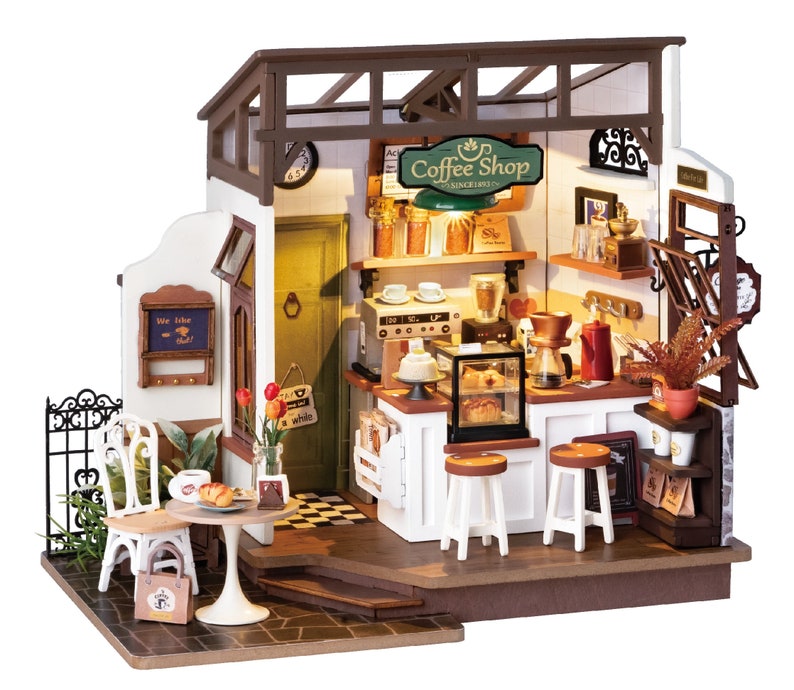 May include: A miniature dollhouse coffee shop with a white exterior, brown roof, and a sign that reads "Coffee Shop Since 1903". The shop has a small patio with a table and chairs, and a window with a sign that reads "We love what we do".
