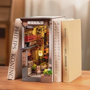 Può includere: Un inserto per libreria in legno che raffigura una scena di strada giapponese in miniatura con una lanterna luminosa, un balcone rosso, dettagli di fiori di ciliegio e una piccola statuetta di gatto. La struttura reca la scritta giapponese ふくろ町 tra due libri.