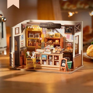 May include: A miniature bakery diorama with a brown exterior, featuring shelves stocked with miniature baked goods, a counter, and a small chalkboard sign. The sign above the shop reads "Bakery & Desserts". The word "OPEN" is visible.