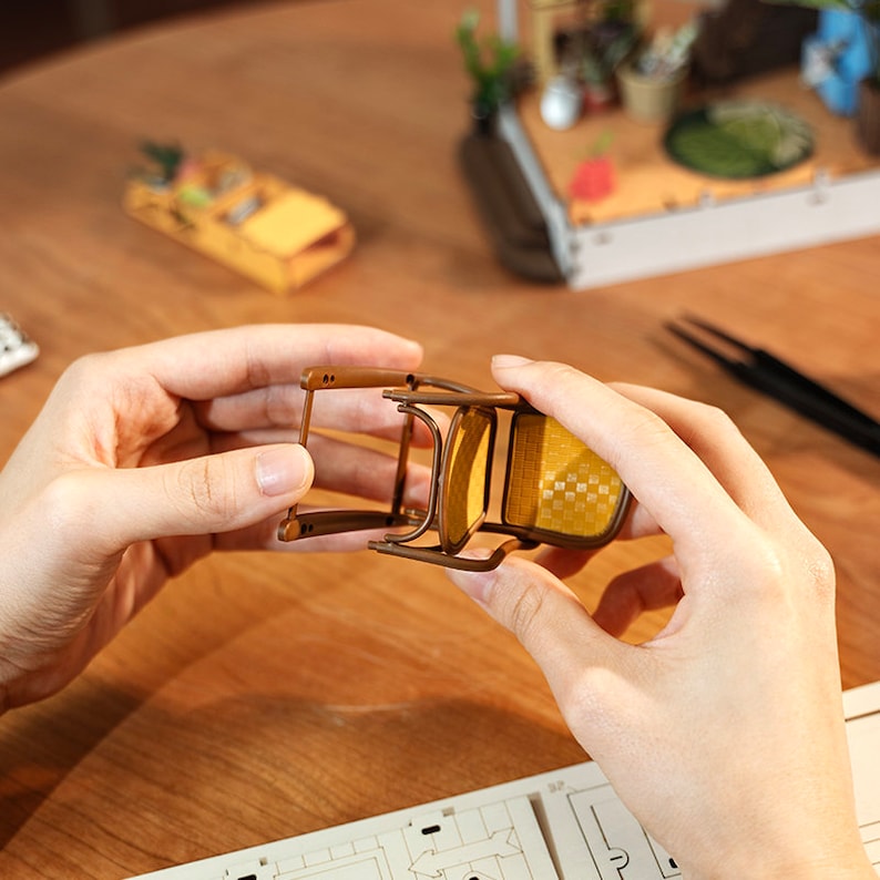 以下が含まれることがあります： 茶色のフレームと籐製の座面を持つミニチュアの木製椅子。椅子は両手で持たれています。