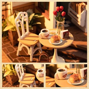 May include: A miniature dollhouse scene with a white table and two chairs, a cup of coffee, a croissant, and a vase of red flowers. The table is set with a plate and a napkin. The scene is set in a cafe or restaurant.