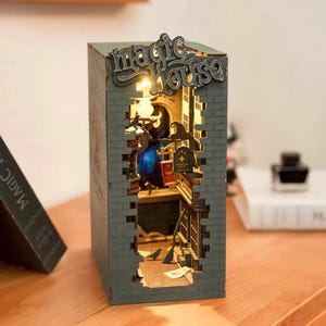 May include: A decorative wooden book nook with a "Magic House" sign. The miniature scene features a witch's hat, a cauldron, and a street scene with birds. The book nook is illuminated with warm light, creating a cozy atmosphere.