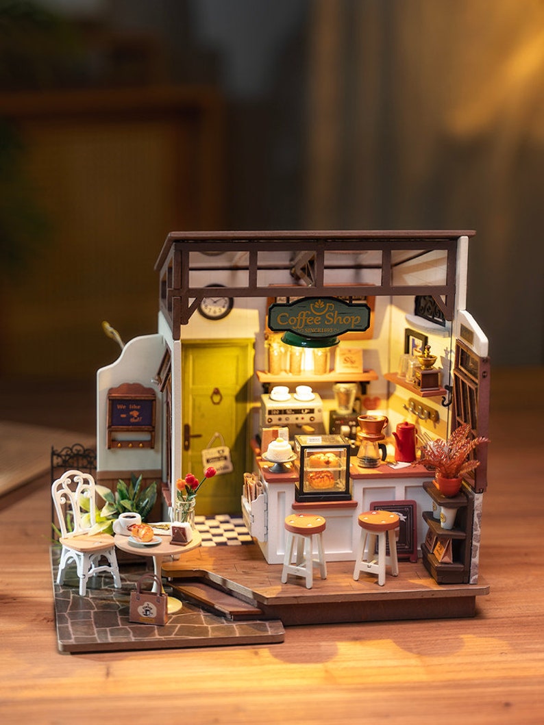 May include: A miniature wooden dollhouse coffee shop with a brown wooden exterior, a green sign that reads "Coffee Shop", and a white interior with a counter, stools, and a table with chairs. The dollhouse is lit up with warm yellow lights.