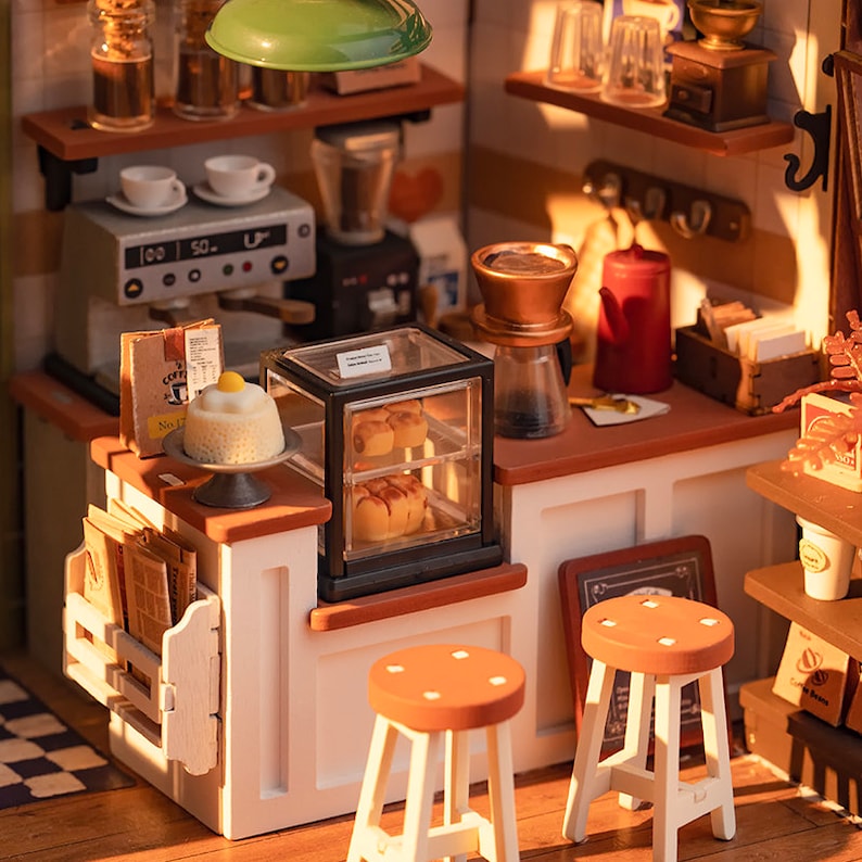 May include: A miniature dollhouse coffee shop with a white counter, two stools, a display case with pastries, a coffee maker, and a sign that says "No Coffee No Life".