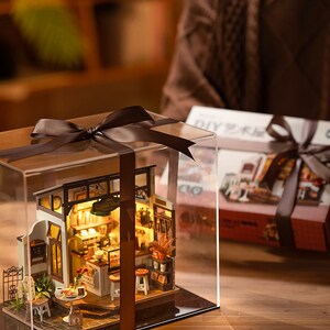 May include: A miniature coffee shop dollhouse kit in a clear plastic display case with a brown ribbon. The dollhouse is lit up and has a sign that says "Coffee Shop".