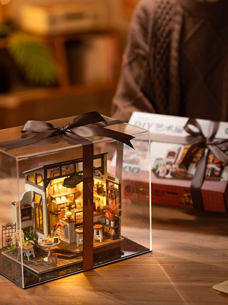 May include: A miniature coffee shop dollhouse kit in a clear plastic display case with a brown ribbon. The dollhouse is lit up and has a sign that says "Coffee Shop".