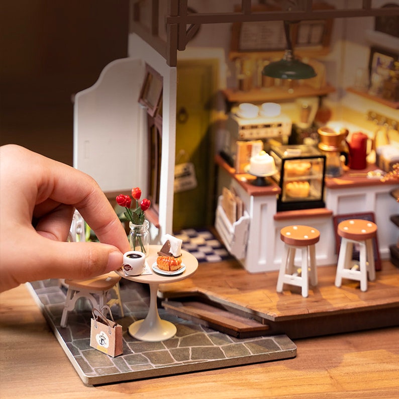 May include: A miniature dollhouse coffee shop with a white counter, brown stools, and a small table with a cup of coffee and a pastry. The dollhouse is made of wood and has a realistic design.