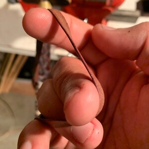 May include: A close-up of a hand holding a thin, brown leather strip. The leather is folded in half and the hand is holding it in place.