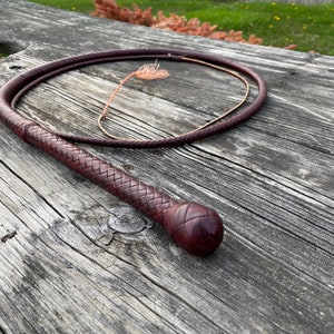 May include: A brown leather bullwhip with a braided handle and a thick, rounded end. The whip is coiled on a wooden surface.