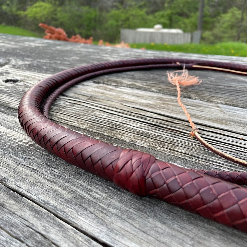 May include: A close-up of a dark red leather braided whip. The whip is lying on a wooden surface. The whip is made of multiple strands of leather that are braided together.