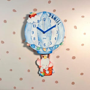 May include: A decorative blue hot air balloon clock with the name "DANIEL" on the clock face. The clock features an elephant in a basket and is decorated with clouds and a crown. The clock hands are blue.