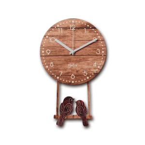 May include: A round wooden wall clock with a light brown wood grain finish. The clock face has white numbers and hands. Below the clock face, two brown bird figures sit on a wooden swing. The clock has the word "Dekeri" printed on the clock face.