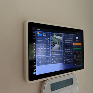 May include: A white tablet mounted on a wall displays a home automation interface with a weather forecast, security camera feeds, and temperature readings. The text on the screen reads "23.1°C", "27°", and "26°".