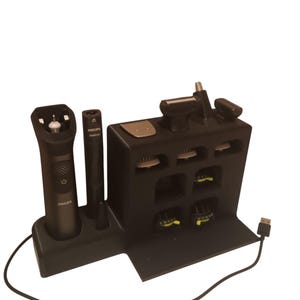 Può includere: Un kit per la cura della persona Philips nero con molteplici accessori, tra cui un rifinitore, un rifinitore per peli del naso e vari pettini. Il kit è esposto su un supporto nero con un cavo di ricarica USB.