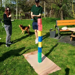 May include: A person playing a game of stacking colorful blocks outdoors. The blocks are stacked in a tower on a wooden base. The person is wearing a black baseball cap and a green shirt. There is a dog in the background.