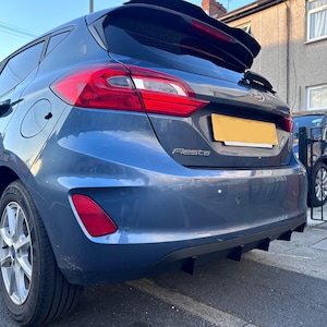 Op de afbeelding: Een blauwe Ford Fiesta hatchback met een achterspoiler en een zwarte diffuser. De auto heeft een kentekenplaat met de tekst "Fiesta" en een achterbumper met een zwarte diffuser.