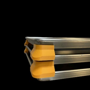 Può includere: Primo piano di un vassoio di metallo con due supporti in plastica giallo-arancio impilati. Il vassoio ha una finitura metallica lucida. I supporti sono rettangolari con bordi arrotondati, che offrono un contrasto di colore e consistenza.
