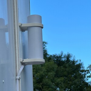 Op de afbeelding: Een witte cilindrische sensor gemonteerd op een witte muur met een witte beugel. De sensor is verbonden met een witte draad. Een groene boom is zichtbaar op de achtergrond.