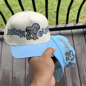 May include: Two baseball caps, one held in hand, featuring a cream-colored crown and a light blue brim. The caps display the text "NORTH CAROLINA" in a stylized font, along with an embroidered ram graphic. The second cap is light blue.
