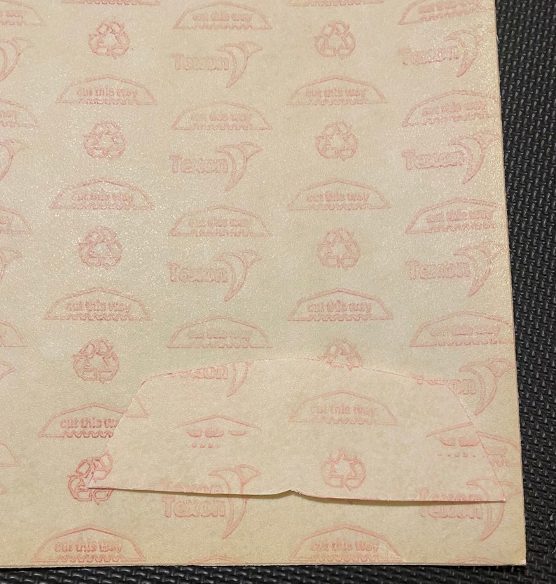 Pode incluir: Um close-up de um papel bege com um padr&atilde;o repetitivo de texto e s&iacute;mbolos rosa. O texto inclui "cut this way" e o nome da marca "Texon". Um s&iacute;mbolo de reciclagem tamb&eacute;m est&aacute; presente. O papel est&aacute; dobrado na parte inferior.