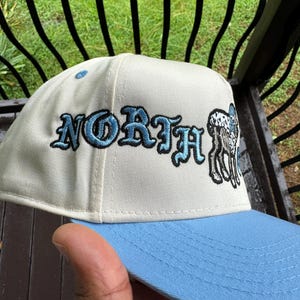 May include: A baseball cap with a light blue brim and a cream-colored crown. The word "NORTH" is embroidered in blue script, with a black and white deer graphic on the side.