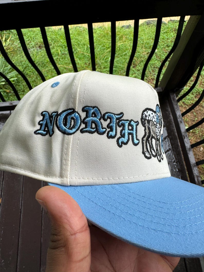 May include: A baseball cap with a light blue brim and a cream-colored crown. The word "NORTH" is embroidered in blue script, with a black and white deer graphic on the side.