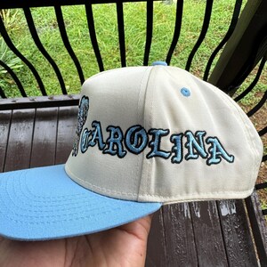 May include: A cream-colored baseball cap with a light blue brim. The word "CAROLINA" is embroidered in light blue with black outlines across the front of the cap. The cap is held by a person.