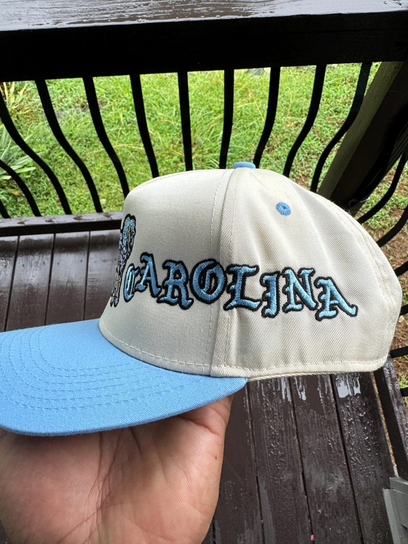 May include: A cream-colored baseball cap with a light blue brim. The word "CAROLINA" is embroidered in light blue with black outlines across the front of the cap. The cap is held by a person.