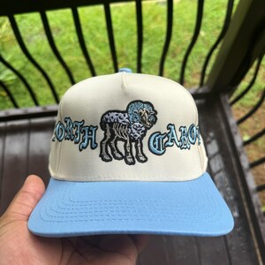 May include: A cream-colored baseball cap with a light blue brim. The cap features the embroidered words "NORTH CAROLINA" in a stylized font, along with an illustration of a ram.