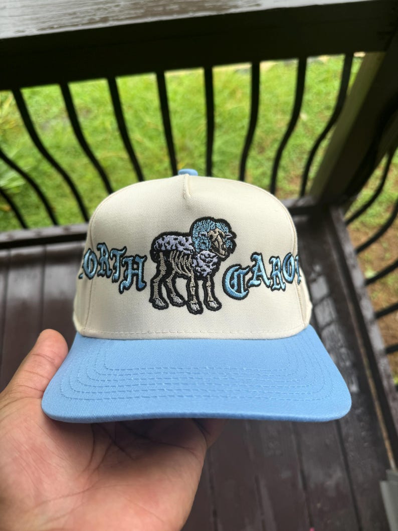 May include: A cream-colored baseball cap with a light blue brim. The cap features the embroidered words "NORTH CAROLINA" in a stylized font, along with an illustration of a ram.
