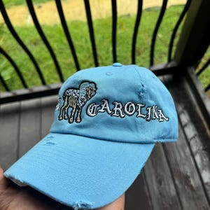 May include: Light blue baseball cap with distressed details. The cap features embroidered text that reads "CAROLINA" in white, along with a ram graphic. The cap is made of a soft fabric and has a curved brim.