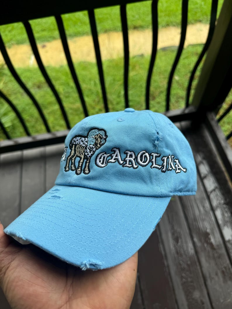 May include: Light blue baseball cap with distressed details. The cap features embroidered text that reads "CAROLINA" in white, along with a ram graphic. The cap is made of a soft fabric and has a curved brim.