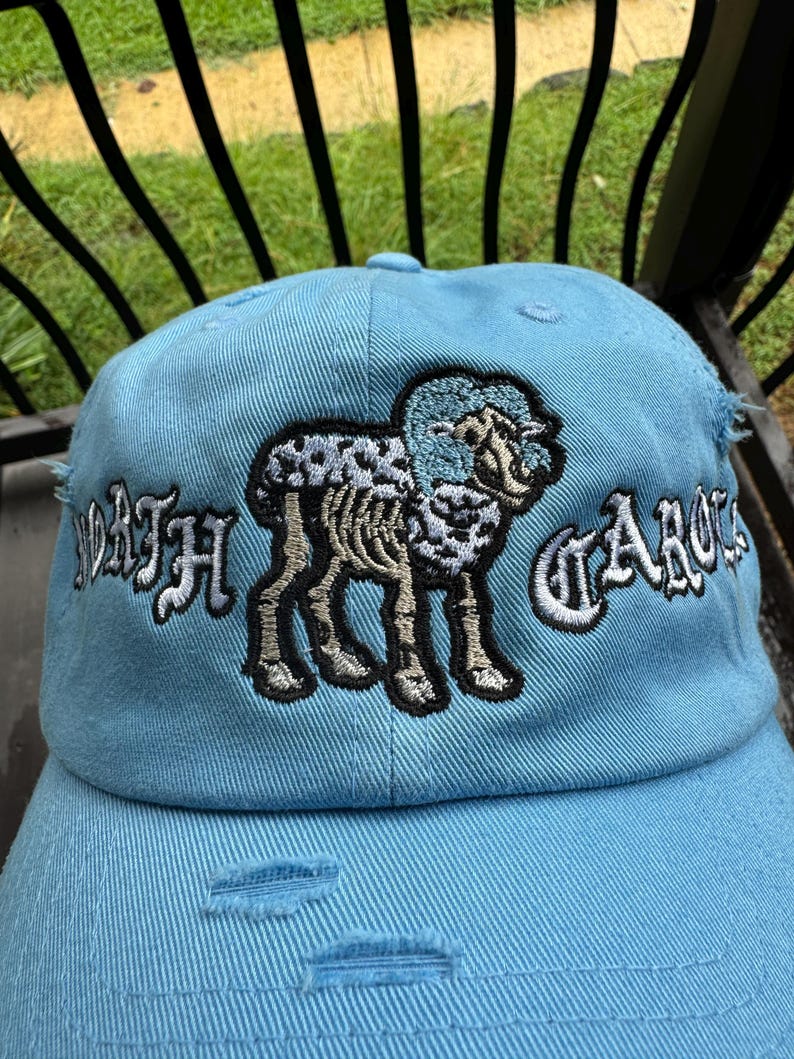 May include: Light blue baseball cap with distressed details. Features an embroidered ram graphic and the words "NORTH CAROLINA" in white. A stylish accessory for fans of the state.
