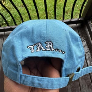 May include: Light blue baseball cap with the word "TAR..." embroidered in white. The cap has a curved brim and an adjustable strap with a metal buckle. The cap is made of a soft, durable fabric.