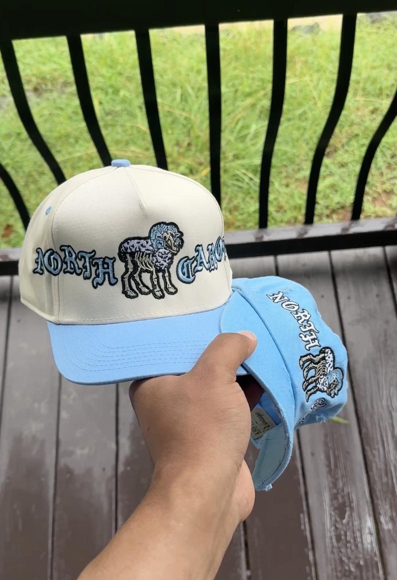 May include: Two baseball caps, one held in hand, featuring a cream-colored crown and a light blue brim. The caps display the text "NORTH CAROLINA" in a stylized font, along with an embroidered ram graphic. The second cap is light blue.