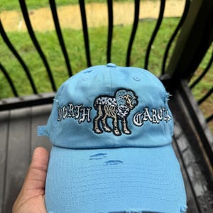 May include: Light blue distressed baseball cap with embroidered text "NORTH CAROLINA" and a ram graphic. The cap has a curved brim and adjustable strap. The hat is a classic accessory.