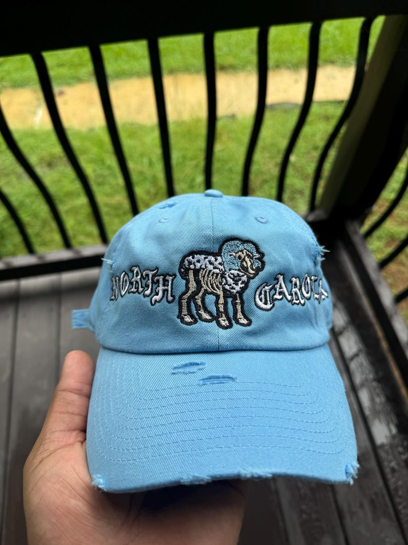 May include: Light blue distressed baseball cap with embroidered text "NORTH CAROLINA" and a ram graphic. The cap has a curved brim and adjustable strap. The hat is a classic accessory.