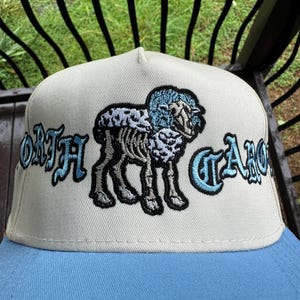 May include: A beige and light blue baseball cap featuring an embroidered design of a sheep with the text "NORTH CAROLINA" in a stylized font. The cap has a light blue brim and a structured crown.