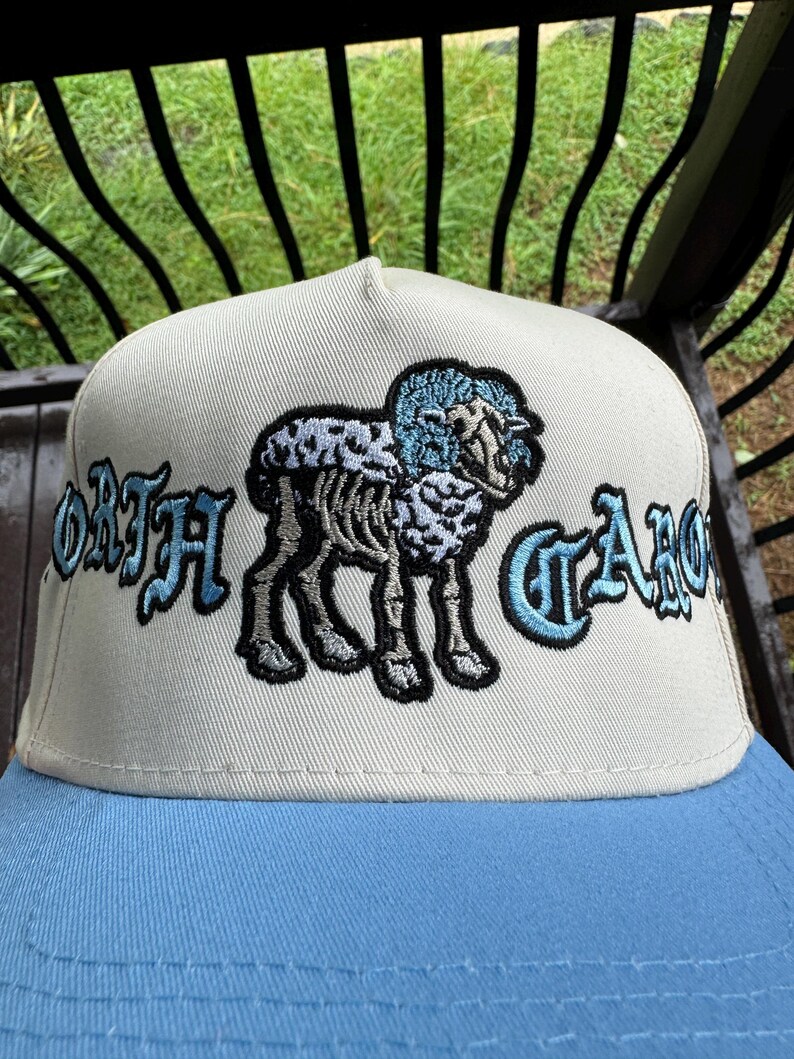 May include: A beige and light blue baseball cap featuring an embroidered design of a sheep with the text "NORTH CAROLINA" in a stylized font. The cap has a light blue brim and a structured crown.