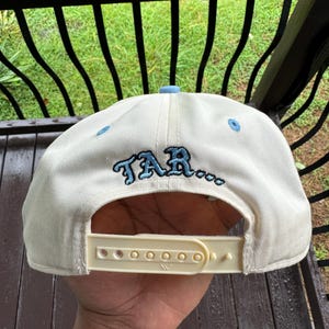 May include: Cream-colored baseball cap with a light blue brim and embroidered text "TAR..." in blue. The cap has a white adjustable snap closure. The cap is made of a durable fabric.